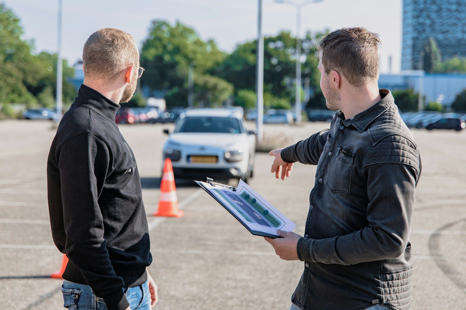 Instructor de manejo con una carpeta explica una prueba en un estacionamiento con conos (foto ilustrativa)